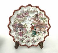 Front view of vintage Japanese scallop-edged ceramic plate with colorful painted Geisha and village scene, red scalloped border, displayed on a wooden stand.