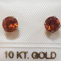 Front view of pair of round faceted orange-red spessartite stud earrings set in 10K yellow gold, displayed on card marked '10 KT. GOLD'.