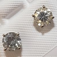 Close-up photo of a pair of 14K white gold stud earrings, each with a round moissanite stone, on a textured white background.