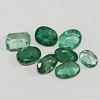 Nine loose emerald gemstones placed on a white background showing variety of cuts and greens.