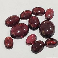 Close-up photo of 10 genuine garnet gemstones showing oval shapes and glossy polished surfaces in deep reddish-brown color.