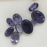 Nine oval cut iolite gemstones in various sizes and blue-violet colors shown on a neutral background.