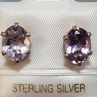 Close-up of pair of oval faceted light purple amethyst earrings set in six-prong sterling silver settings on a white card marked 'STERLING SILVER'.
