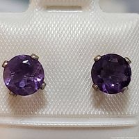 Close-up of two round faceted amethyst stud earrings with sterling silver prong settings on a white display.