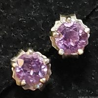 Close-up image of a pair of stud earrings each with a round faceted purple amethyst stone set in sterling silver prongs.