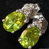 Close-up of the pair of sterling silver earrings with faceted oval light green peridot gemstones and small round clear stones above them, showing front and slight angle views.