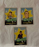 Front view of three 1967-68 O-Pee-Chee NHL Second Team hockey cards for Johnny Bucyk, Ed Giacomin and Rod Gilbert, showing player portraits and star background with banner.