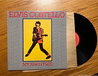 Front cover of Elvis Costello My Aim Is True vinyl record showing checkered black and white border and image of Costello in a red jacket.
