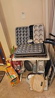 Photo of several metal muffin pans/trays stacked along a small lap tray table, with oven mitts and some kitchen utensils below.