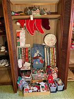 Closet shelf and floor with large containers and boxes holding rolls of Christmas wrapping paper, gift bags, various ribbons and bows, four red Christmas stockings hanging, a Santa hat, and a ceramic house with internal light surrounded by small decorative trees.