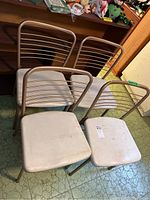 Four beige metal-framed folding chairs with white vinyl seats, arranged facing away. Chairs show moderate wear and dirt marks on seats.