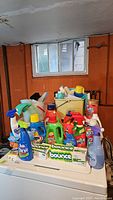 Lot of laundry and cleaning products placed on a white surface with a background of a basement wall and window. Includes detergents, sprays, one yellow bucket, and gloves.