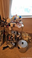 Full view of the kitchenware lot including pots, pans, sponges, table linen stacked on and beside the TV table near the window.