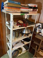 Cookbooks stacked on top shelf with various kitchen items including pans and a mandolin on lower shelves of a metal rack.