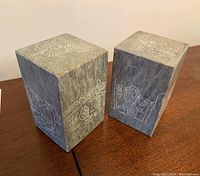Both soapstone bookends positioned on a wooden surface showing engraved butterflies on top and nature/cultural scenes on the sides.