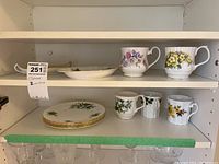 Two shelves showing a total of eight floral patterned mugs, plates, and a small platter, with gold trim visible on plates and some staining noted on mug interiors.