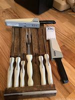 Six white handled Wear Ever cutlery pieces in a wooden rack plus two flatware pieces with black and gray handles stored on floor