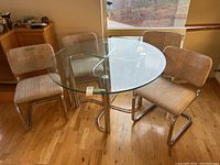 Round glass-top dining table with stainless steel windmill base seen with four upholstered chairs nearby, shown on hardwood floor near window.