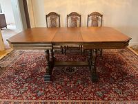 Front view of rectangular dining table with wooden veneer top and two wood pedestal legs on trestle base, surrounded by chairs and placed on area rug.