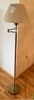 Front side view of the brass swing arm floor lamp with beige pleated lampshade, shown standing on hardwood floor next to white wall.