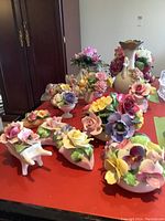Multiple ceramic floral pieces arranged on a table with a wooden cabinet in background. Assortment of ceramic flowers in pastel colors with visible detailing.