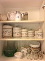 Shelves with cream and floral patterned ceramic canisters of various sizes, matching soap dispenser and salt & pepper shakers, cream colored tea pots, clear glass bowls and plates with stacked cups, shot glasses, and a small ceramic creamer pitcher.