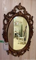 Full view of the oval mirror hanging on a wall, showcasing the ornate gold composite frame with scrolling detail.