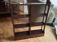 Front view of wood shelf unit showing three shelves and two drawers with metal knobs, placed on hardwood floor near a green upholstered chair.