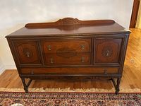 Front view of brown wooden sideboard showing two cupboards and three drawers with brass hardware, decorative carved backsplash on top.