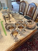 Silver plate goblets, platters, covered serving dishes, glass inserts on a table with two wooden chairs in the background