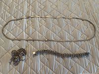 Three items laid out on a quilted surface: silver chain necklace, linked bracelet, and decorative brooch.