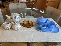 Two tea sets and glass cream and sugar set displayed on white cloth on glass table. Royal Cauldon set includes creamer, sugar bowl, teapot, and platter with floral pattern and gold accents. Glass cream and sugar set has ribbed amber glass cups. Ellgreave blue matte tea set includes teapot, creamer, cup, and matching tray.