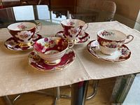 Four teacups and matching saucers including two Queen Anne Lady Eleanor, one Royal Standard, and one Paragon, arranged on cloth on glass table.