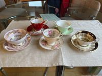 Five porcelain teacups and saucers displayed on a tablecloth showing various floral and gold designs.