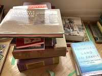 Books stacked on a wooden floor including 'To Serve With Honour', 'My Promised Land' by Ari Shavit, and other reference books along with a cardboard box.