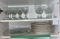 Two shelves showing approximately 17 clear glass wine glasses and stacked plates plus two platters on lower shelf, one decorated with floral design