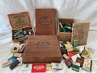 Three wooden cigar boxes arranged open and closed on a white cloth background with assorted disposable lighters and matchbooks surrounding them