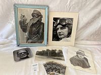 Framed vintage aviator photos, metal tray with aviator image, black and white photo prints, and postcards arranged on a white cloth background.