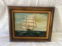 Framed oil painting showing a large sailing ship on the ocean with full white sails under a cloudy sky, set in a wooden frame with subtle texture.