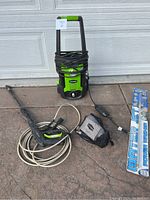 Complete green and black Greenworks pressure washer with power cord coiled on the handle, attached spray gun with beige hose, and additional packaged accessory laid on ground near a garage door.