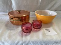 Copper pot with lid, yellow and white Pyrex mixing bowl, two small red glass cups photographed from the front on white cloth background.