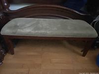 Front angled view of wooden bench with sage green cushion on a light wood floor, showing slight fabric discoloration and wear on wood finish.