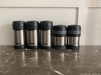 Front view of five stainless steel Thermos containers with black lids and bases, arranged side-by-side on a marble surface against a white background.