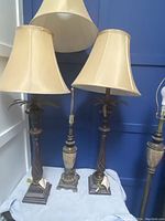 Front view of three lamps on a white surface against a blue wall, including two palm tree style table lamps with cream shades and one additional lamp behind them.