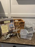 Photo of assorted home decor and organization items including white 'Believe' metal decor, wicker basket, scrapbook page holder, and white wire baskets on a marble surface against white paneled wall.