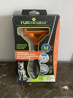 Front view of Furminator undercoat deshedding tool in box showing packaging, brand, and size M for medium dogs.