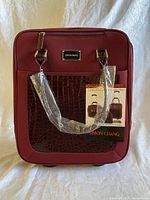 Front view of the maroon rolling carryon bag with faux crocodile texture pocket and wrapped handles, Simon Chang brand tag attached.