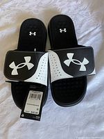 Pair of new black and white Under Armour men's sandals, size 10, with original price tag attached showing a retail price of $40.