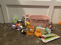 Photo showing pink basket with assorted small plush toys, figurines, candy, wearable items, and small accessories arranged in front of a white paneled wall.