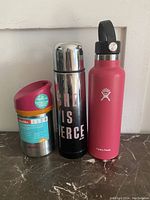 Three stainless steel bottles: Aladdin food jar, black water bottle with text, pink Hydro Flask bottle